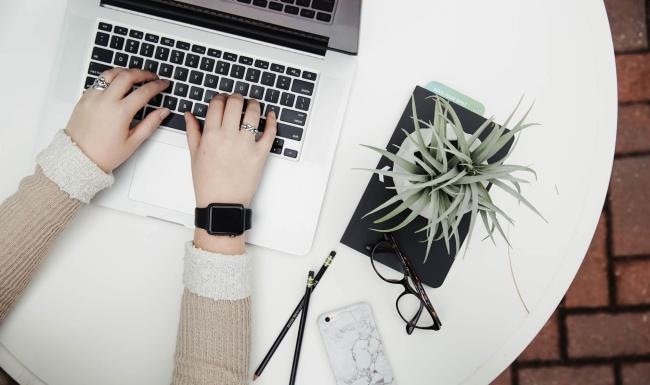overhead shot of woman working on a laptop at a round table with pencils, her cell phone, glasses, notebook and a potted plant